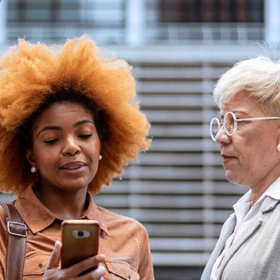 Two women in formal business clothing discussing something seen on a mobile device while standing outside.