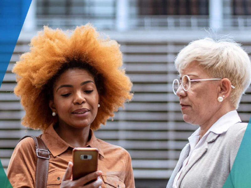 Two women in formal business clothing discussing something seen on a mobile device while standing outside.