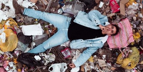 A young male makes a "shush" gesture with his hand while lying on top of a landfill site to convey silence about the environment.