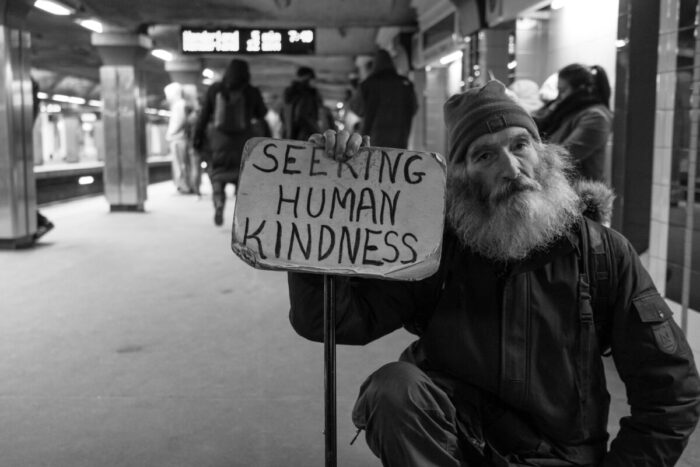 Photo by Matt Collamer on Unsplash. Photo depicts a homeless man in a New York city subway station with a sign saying, "Seeking human kindness."