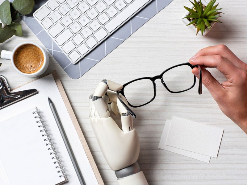 the image show a computer desk, with a cup of coffee and a note book on it. Also show a reading glasses that of it's handle is being hold by a human hand and the other one by a robot hand