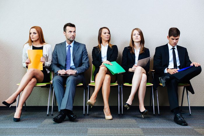 the image shows 5 white people, 2 men and three women, siting on chairs likely to be waiting for job interview