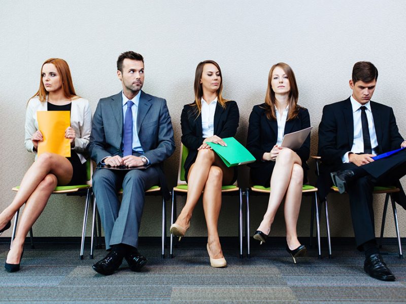 the image shows 5 white people, 2 men and three women, siting on chairs likely to be waiting for job interview