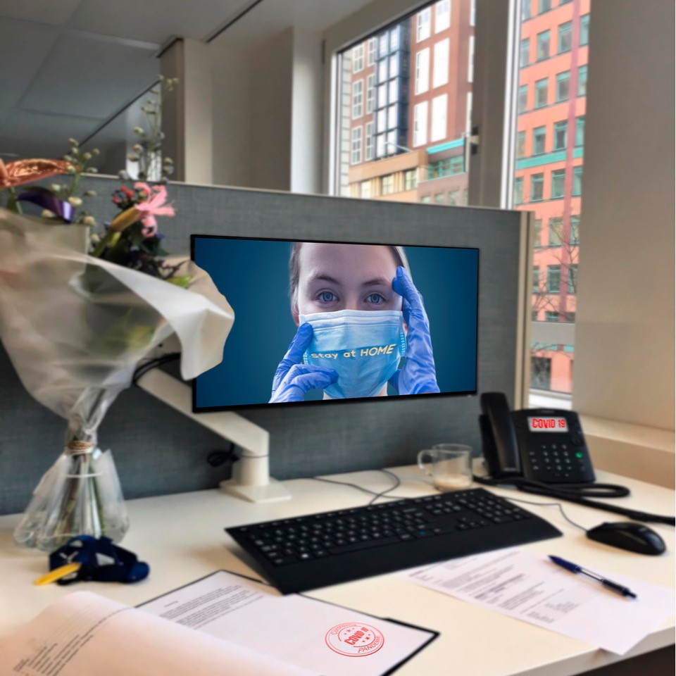 A photo of an office desk with an image on the computer monitor of a nurse in a mask and the phrase, "stay at home" across her mask.